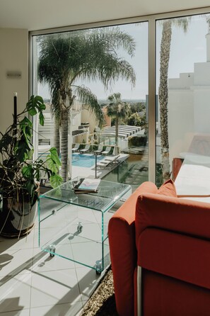 A modern living room with a glass coffee table, a red sofa, and a view of a pool and palm trees. - Hotel Sao Sebastiao de Boliqueime (Loulé)