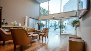 A modern hotel lobby with a wooden floor, leather chairs, a reception desk, and large windows offering a view of the street. - Hotel Sao Sebastiao de Boliqueime (Loulé)