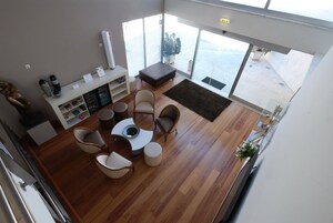 Una sala moderna con una mesa de centro redonda, sillas blancas y marrones, un mueble blanco y un ventanal con vistas al mar.