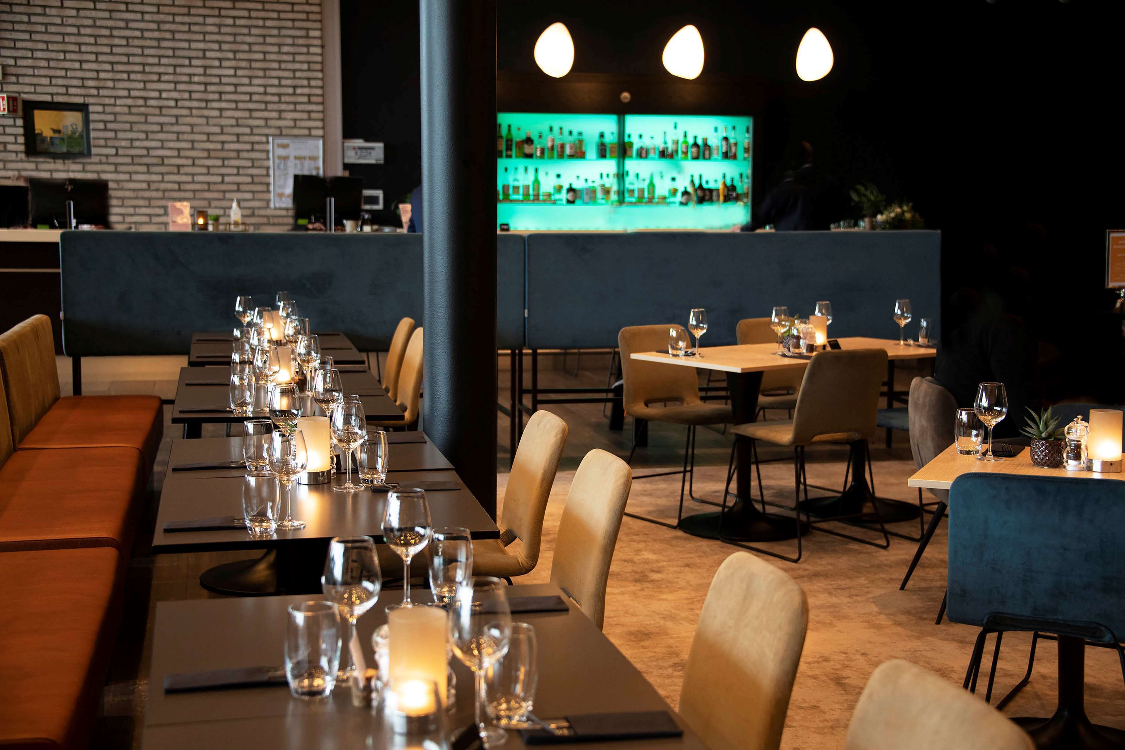 A restaurant with tables set for dining, a bar area with a green-lit display, and a brick wall in the background.