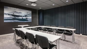 Un salón de conferencias con una larga mesa, sillas, una pantalla de proyector y una pared oscura con líneas verticales.