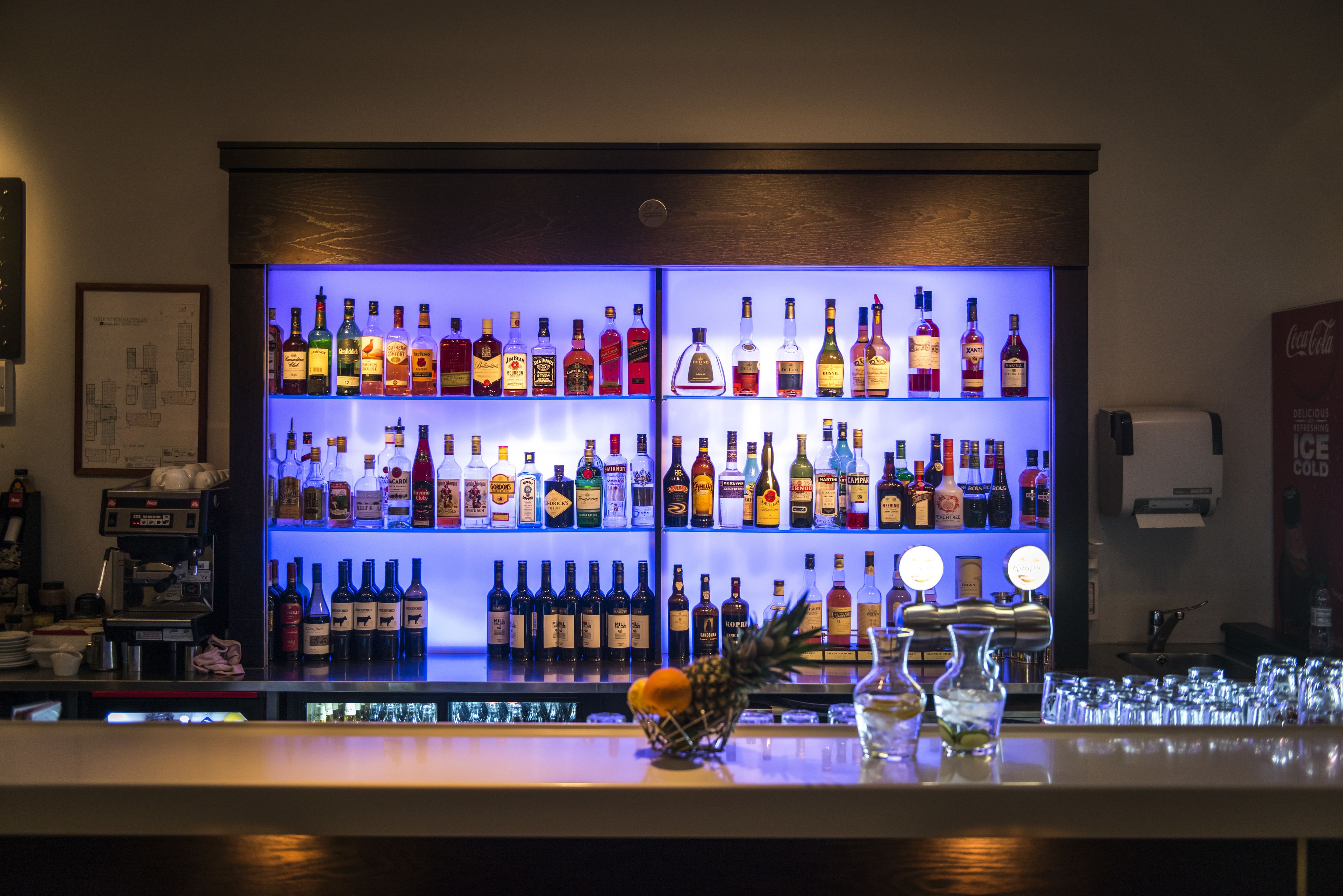 Una zona de bar con estantes llenos de botellas de licores, una piña y vasos sobre el mostrador.