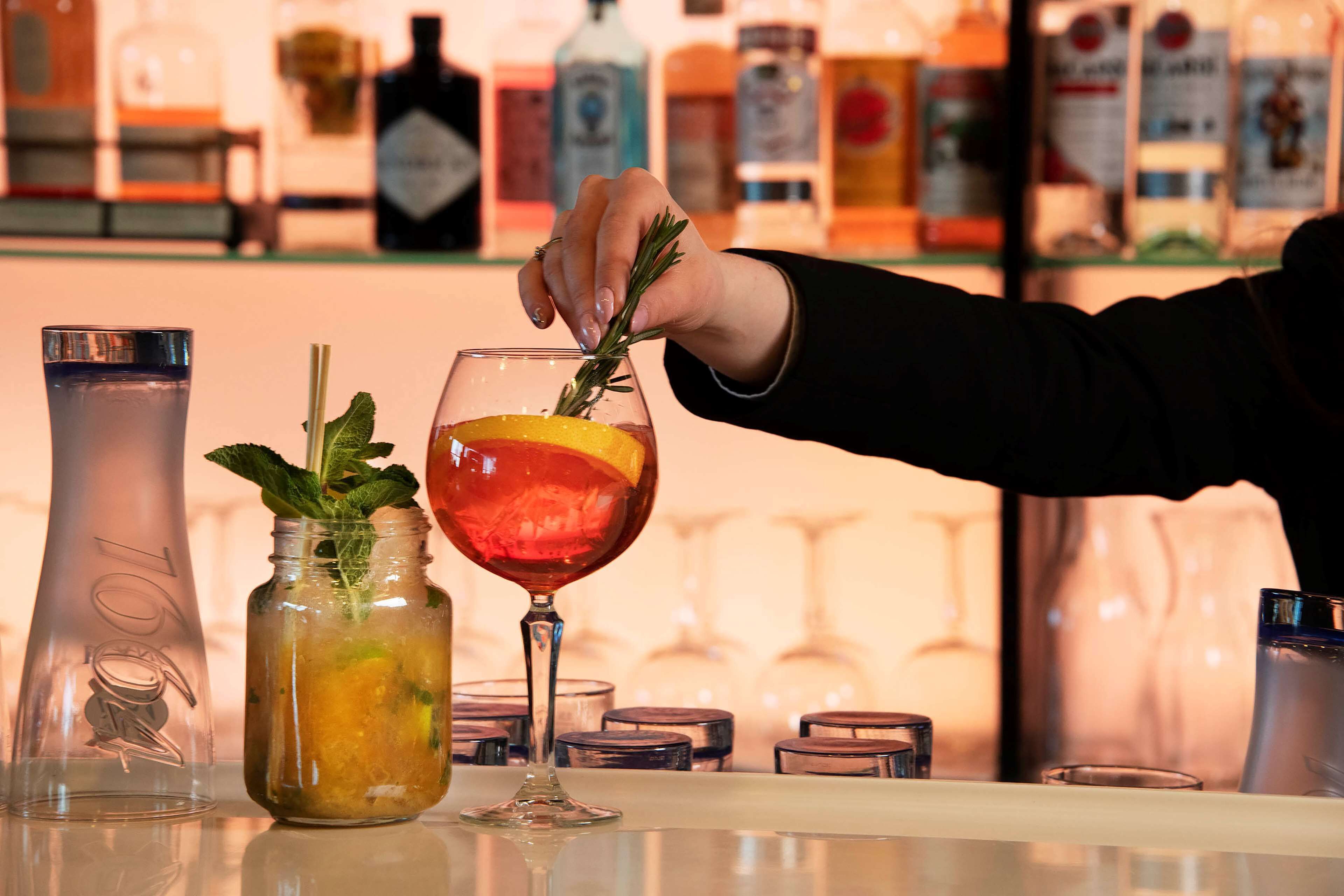 A bar setting with a bartender preparing a drink, a mason jar with a mint garnish, and various bottles in the background.