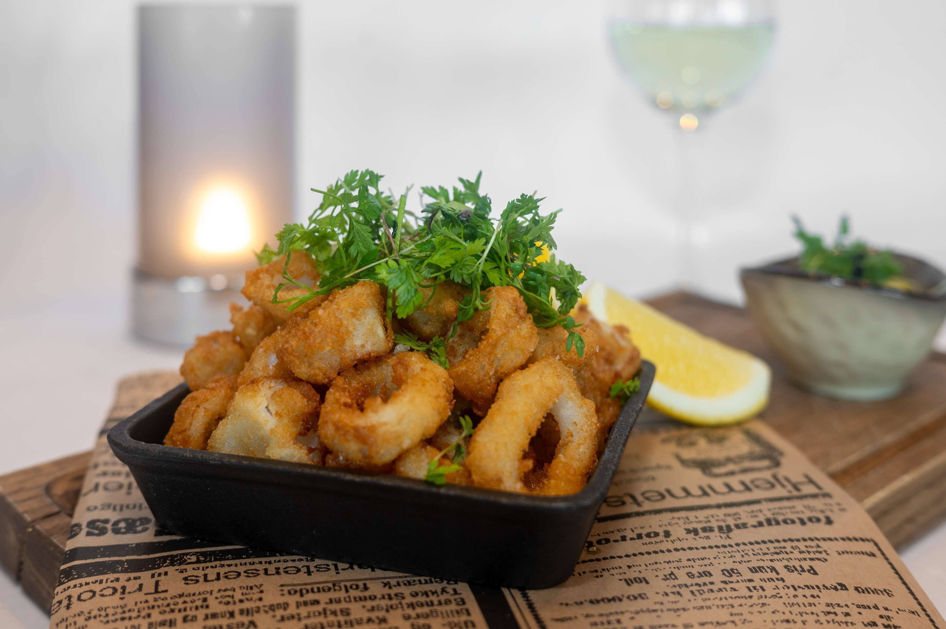 Una bandeja de calamares fritos adornada con perejil, acompañada de un gajo de limón y una copa de vino blanco, sobre una mesa con un periódico debajo.