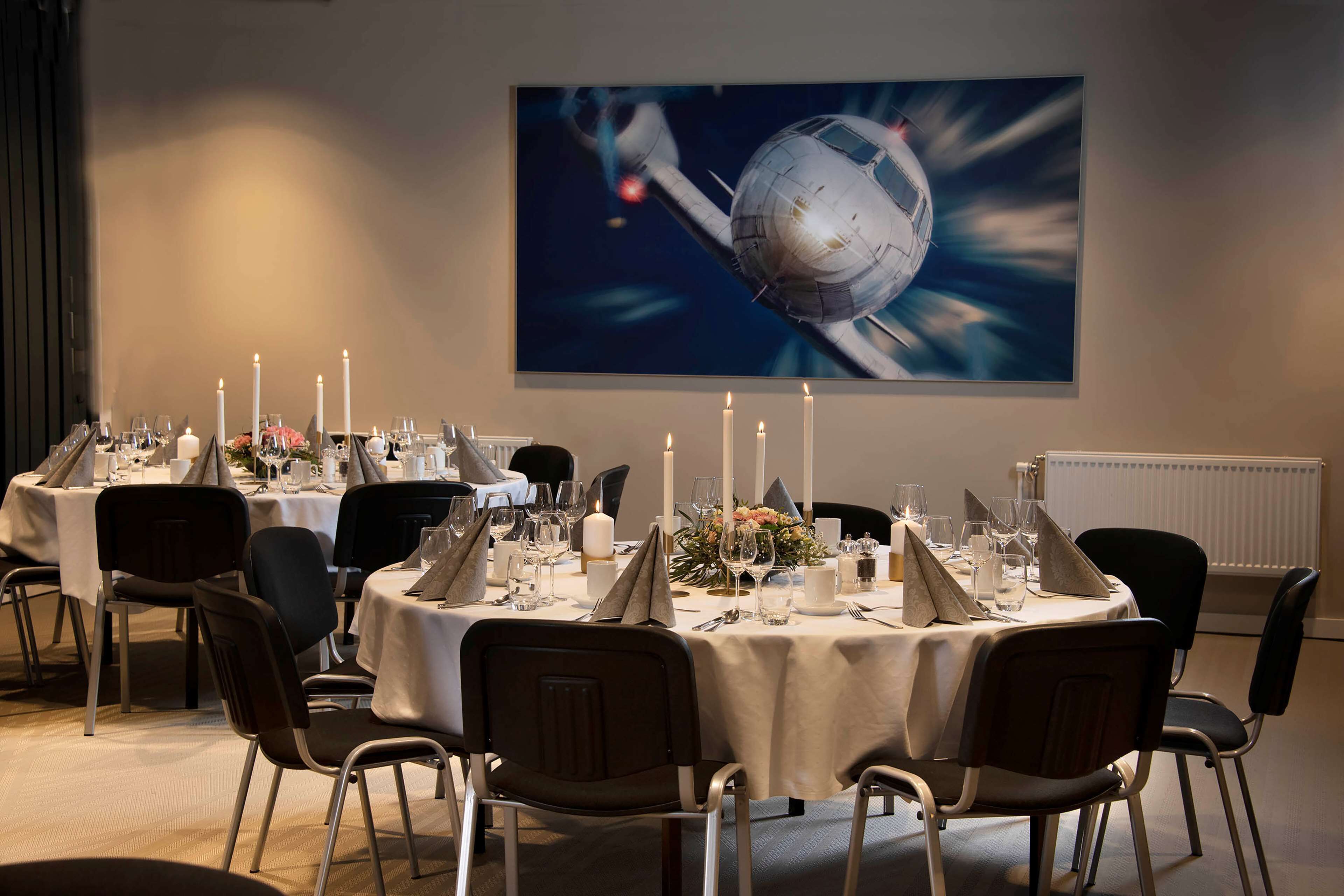 A dining area with round tables set for a meal, featuring white tablecloths, candles, and a large painting of a space scene on the wall.