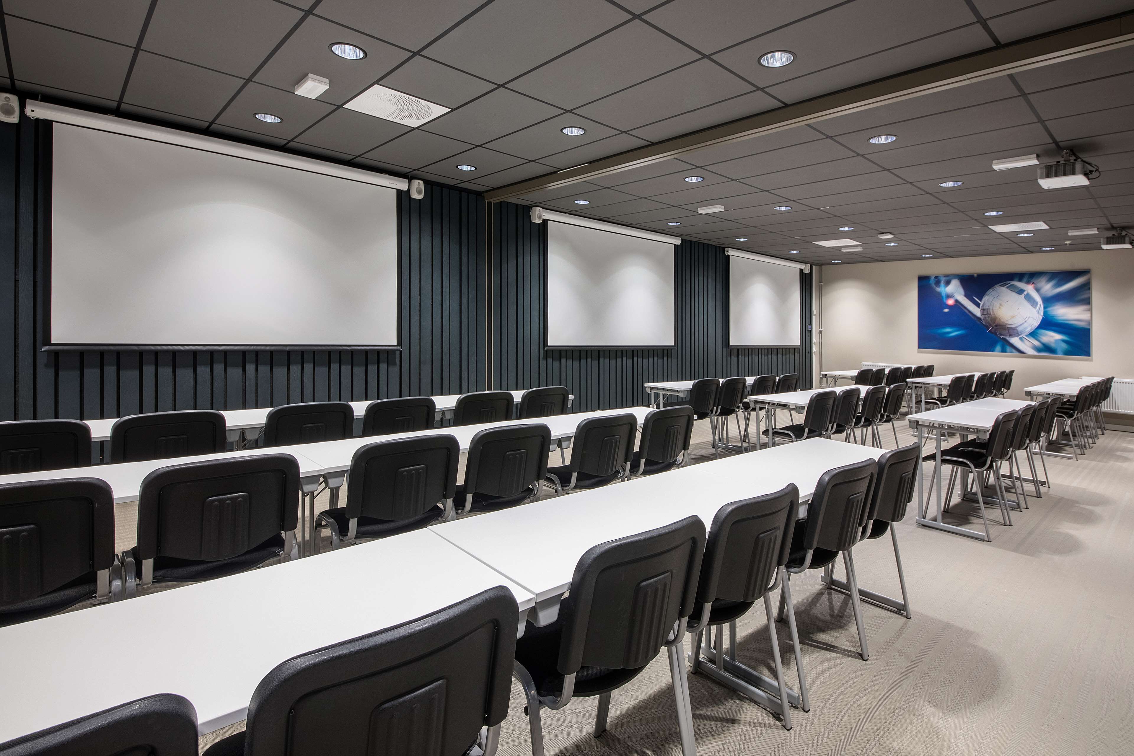 A conference room with long tables, black chairs, a large screen, and a projector.