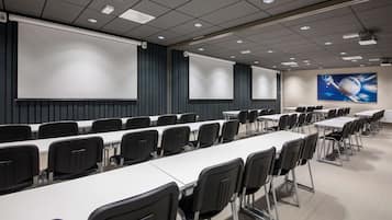 Un salón de conferencias con largas mesas, sillas negras, una pantalla grande y un proyector.