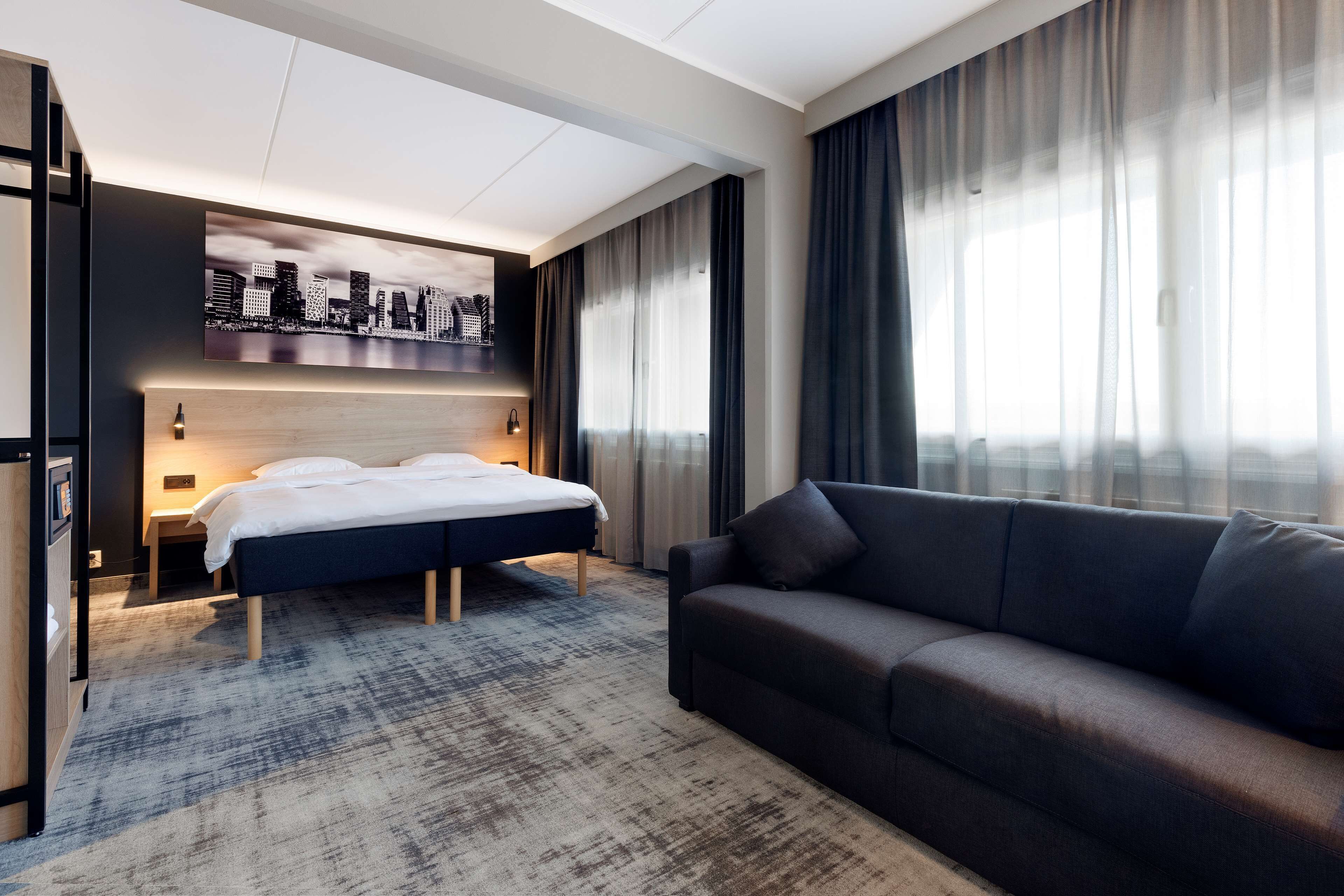 A modern hotel room with a large bed, a sofa, a cityscape artwork, and a city view through the window.