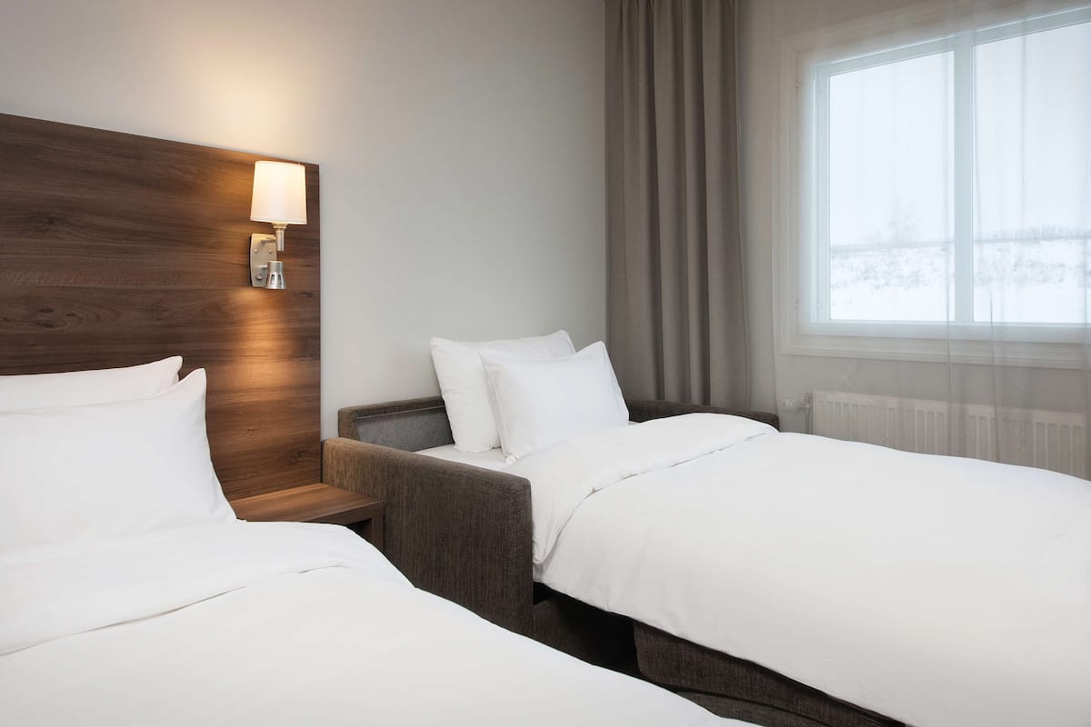 Habitación de hotel con dos camas, cabecero de madera, mesita de noche, lámpara y ventana con cortinas.