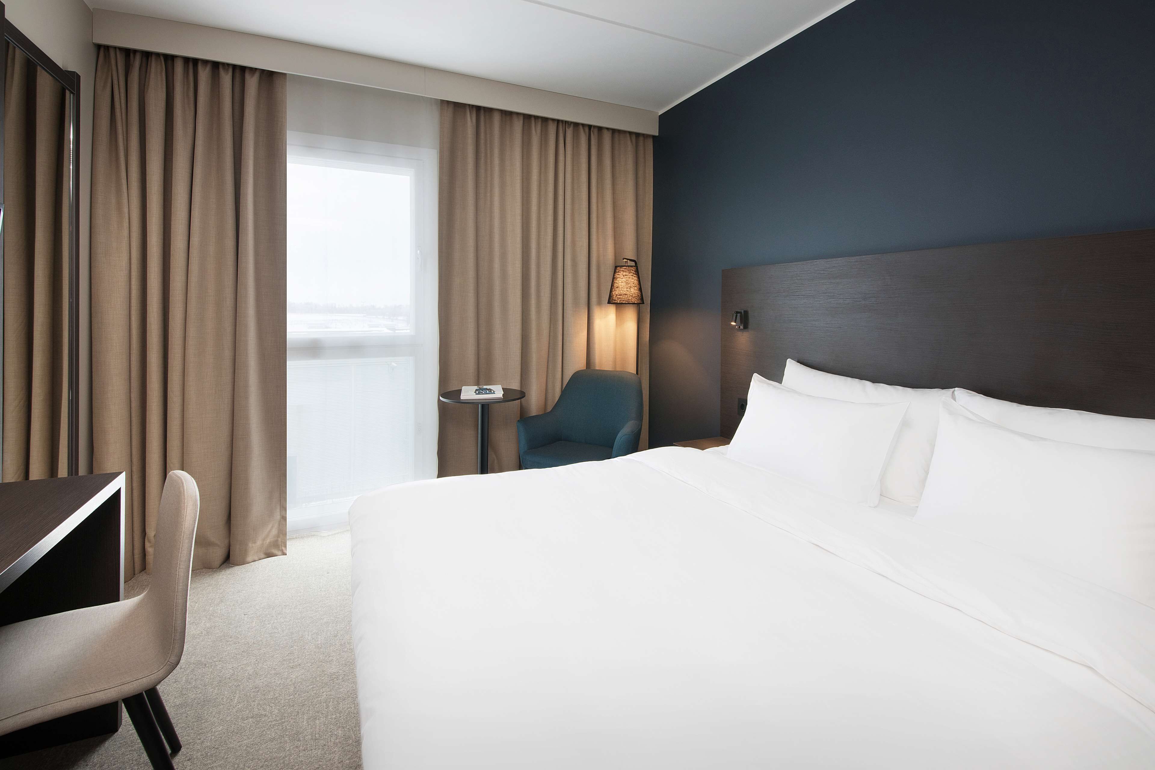 Una habitación de hotel con cama, escritorio, silla, lámpara y vistas al exterior.