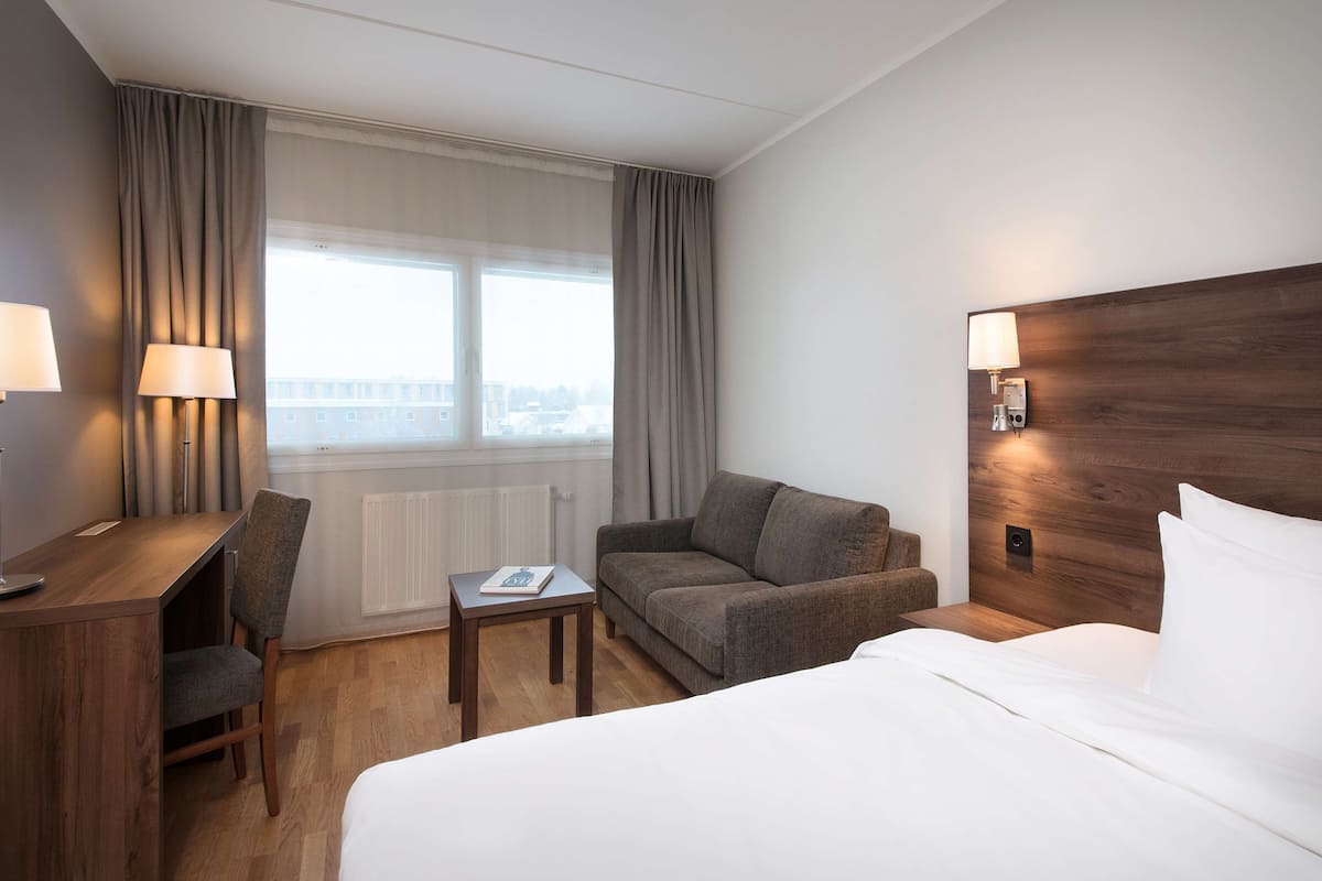 Habitación de hotel con cama, escritorio, silla, mesita, sofá y ventana con cortinas.
