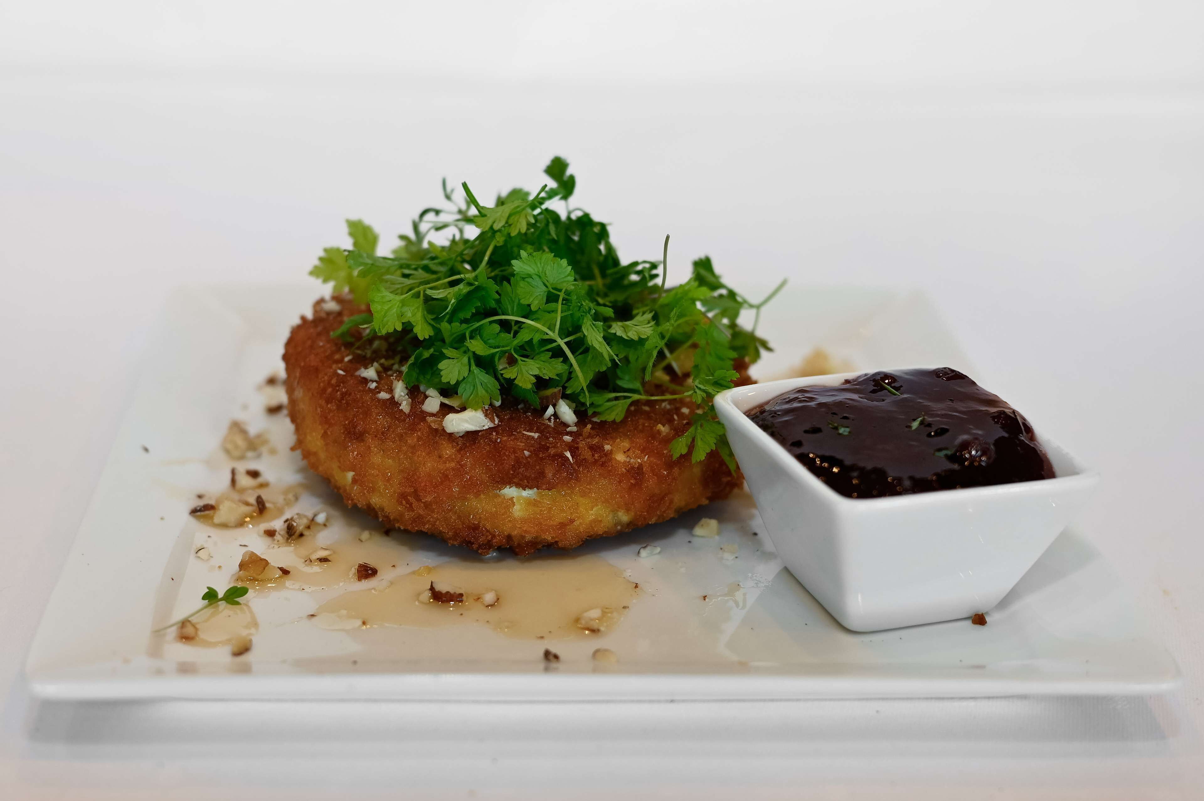 Un plato con un guiso dorado y crujiente, decorado con hierbas frescas y nueces, acompañado de un recipiente pequeño con salsa oscura.