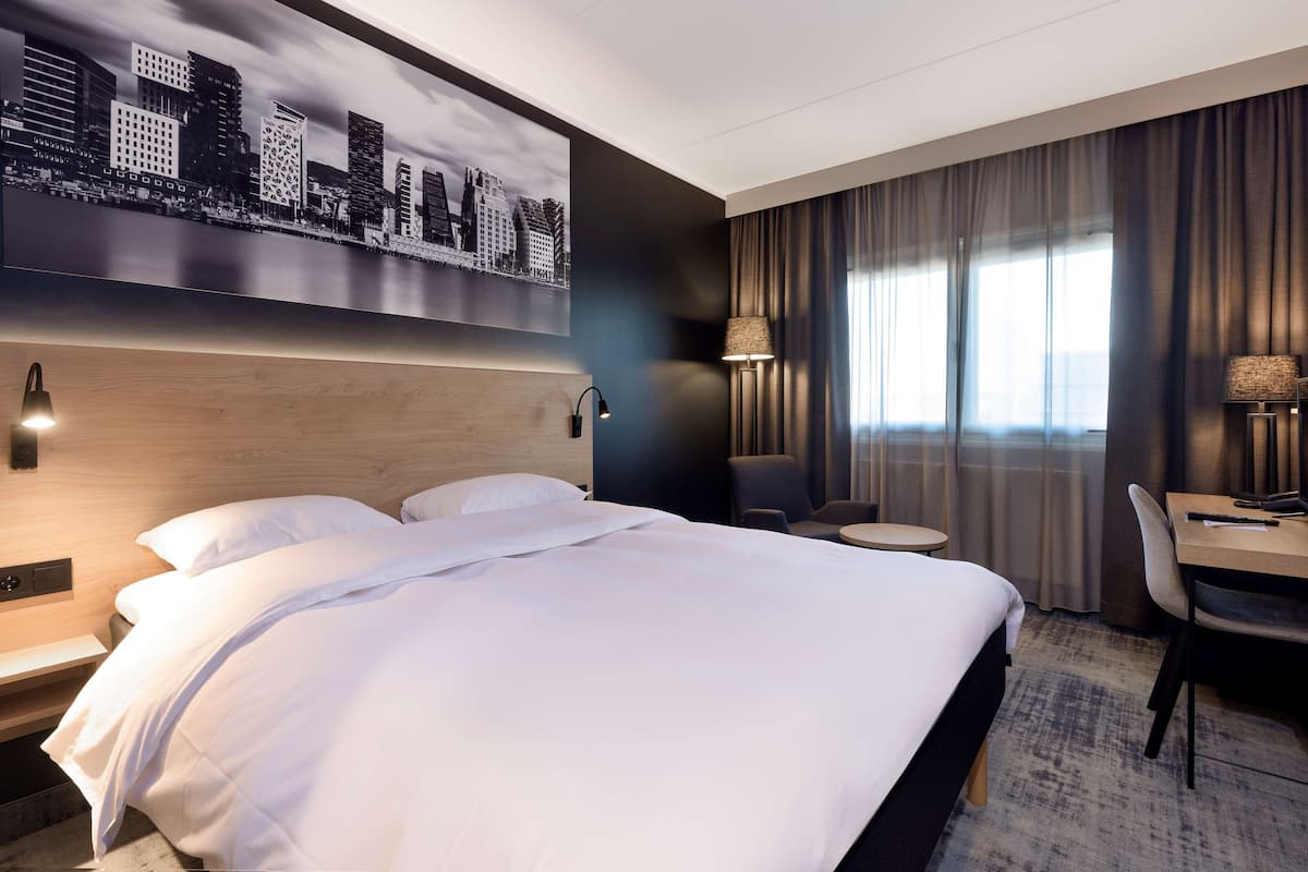 Habitación de hotel con una cama grande, un escritorio, una silla, una lámpara y un cuadro de un paisaje urbano en la pared.
