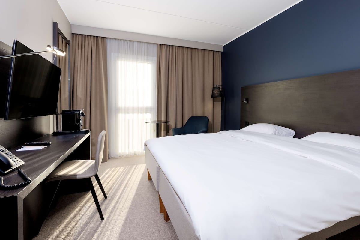 Habitación de hotel con cama, escritorio con computadora, silla, televisor y ventana con cortinas.