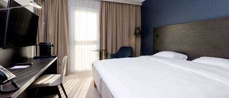 Une chambre d’hôtel avec un lit, un bureau avec un ordinateur, une chaise, une télévision et une fenêtre avec des rideaux.