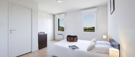 Habitación de hotel con cama, mesita de noche, lámpara y vistas al exterior a través de una ventana.