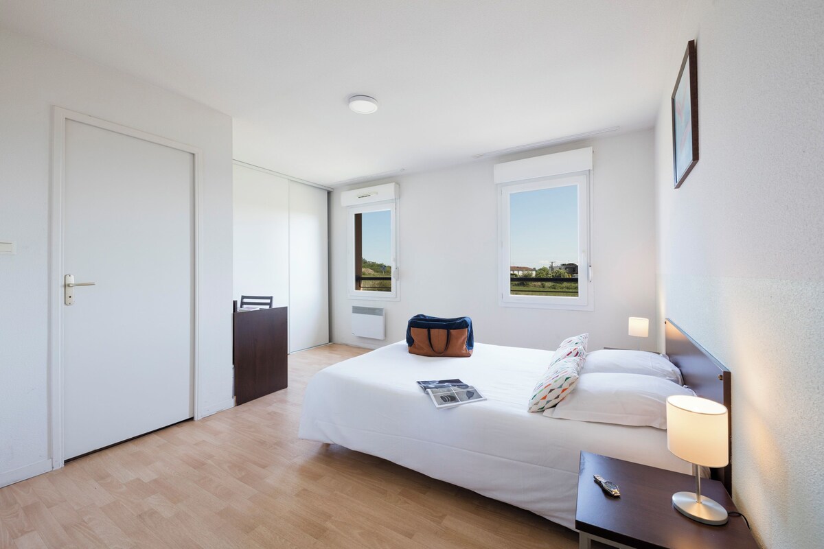 A hotel room with a bed, bedside table, lamp, and a view of the outdoors through a window.