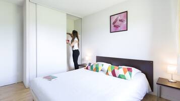 Hotellihuone, jossa on sänky, yöpöytä, lamppu, peili ja taulu seinällä.
