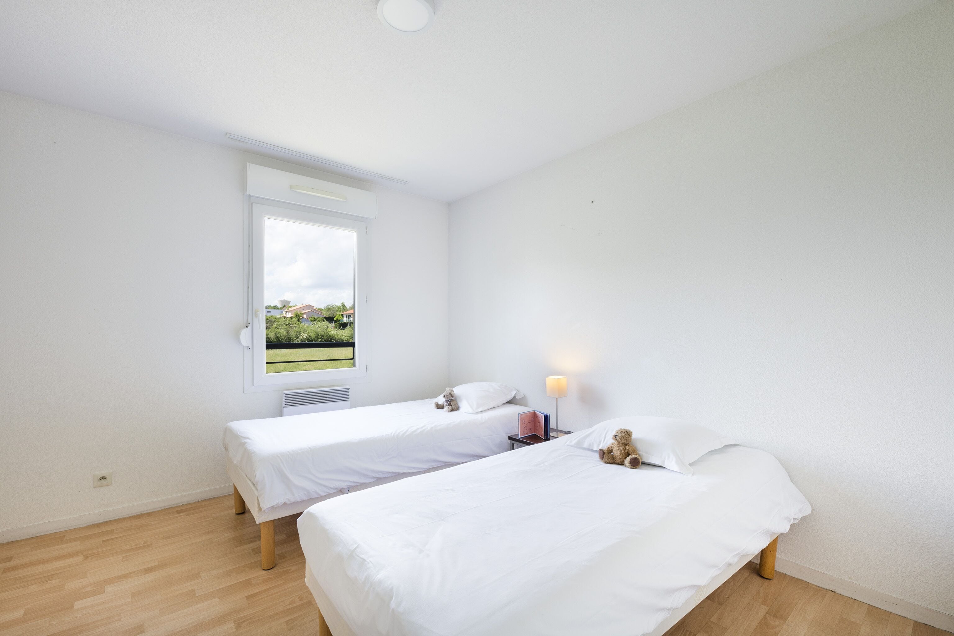 Een kamer met twee eenpersoonsbedden, een raam met uitzicht op groen, een bedlampje en een klein tafeltje met een boek en een teddybeer.