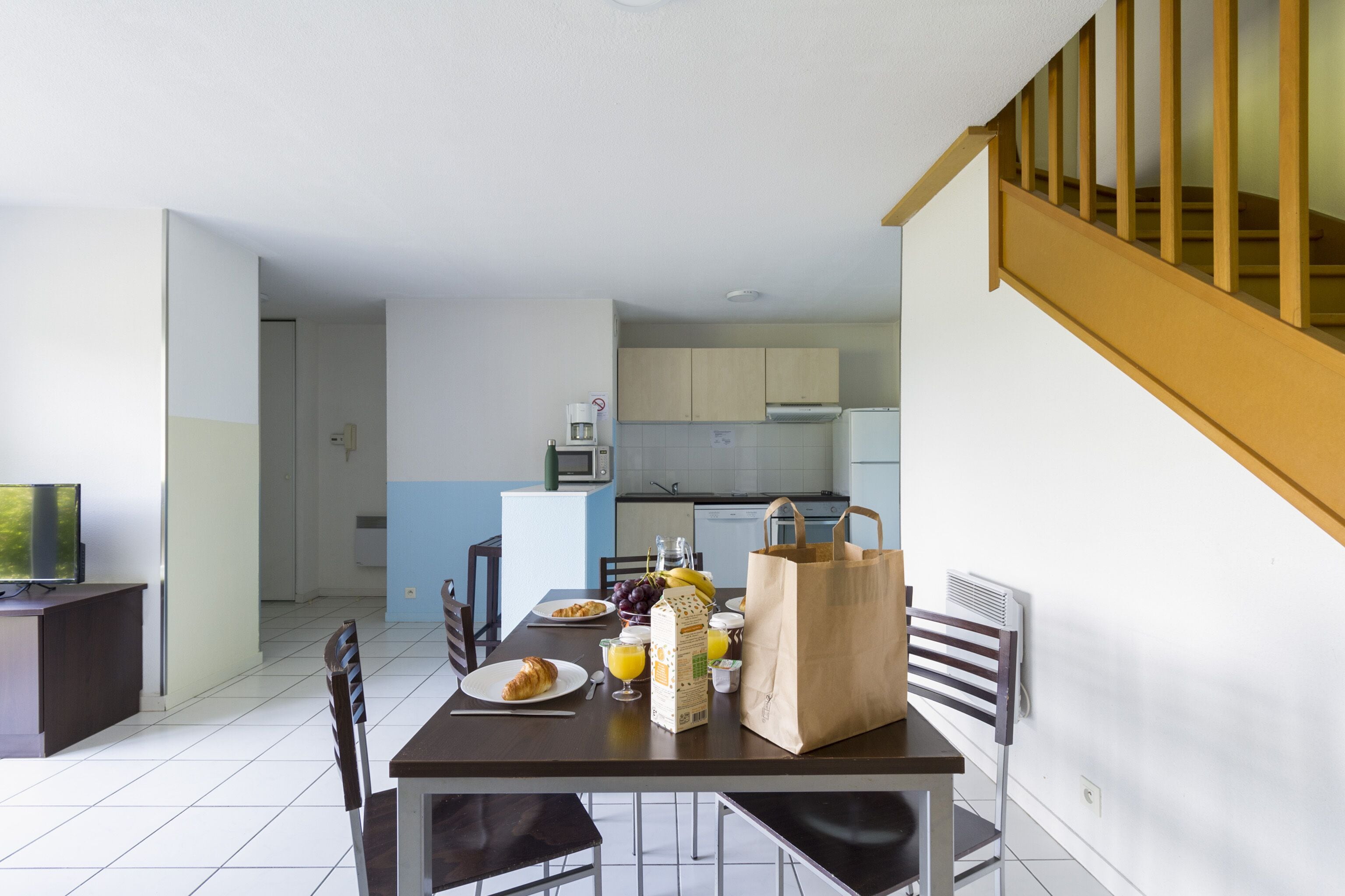 A dining area with a table set for breakfast, including croissants, fruit, and juice. There is a bag of groceries and a microwave in the background.