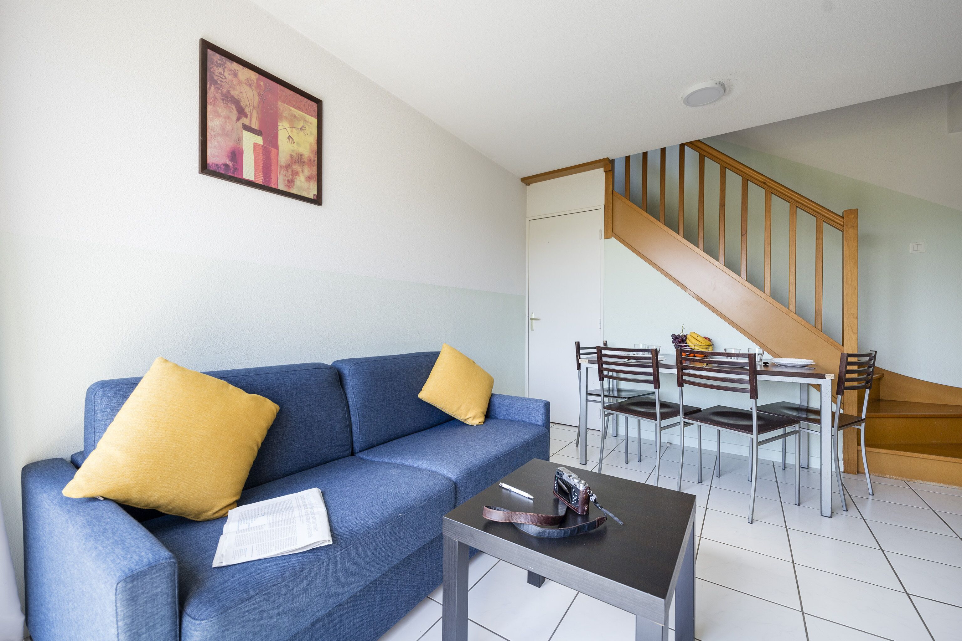 Una sala de estar con un sofá azul, cojines amarillos, una mesita y una escalera que lleva a un comedor con sillas y una mesa.
