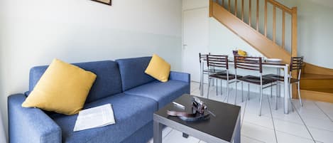 Una sala de estar con un sofá azul, cojines amarillos, una mesita y una escalera que lleva a un comedor con sillas y una mesa.