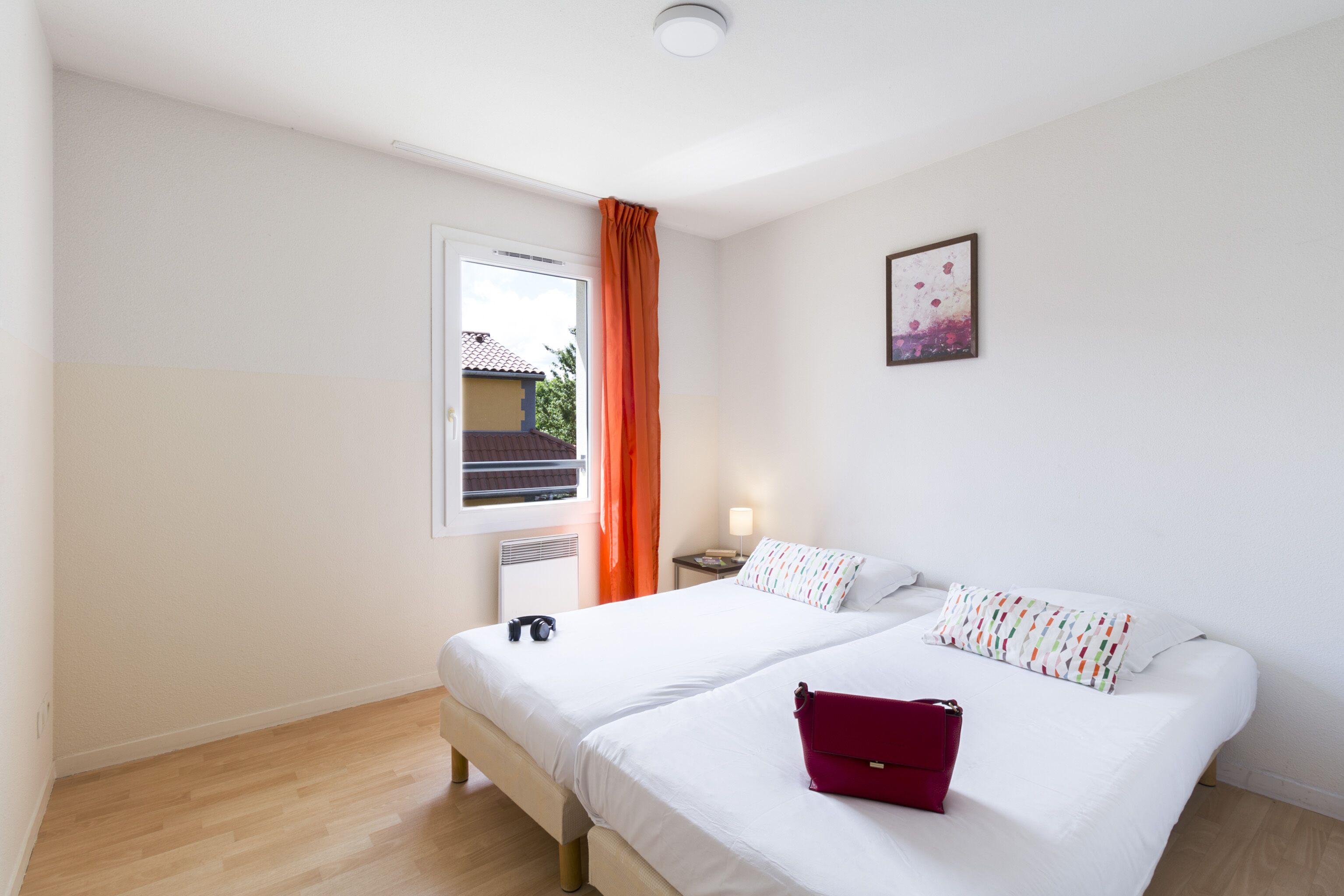 Habitación de hotel con dos camas, una ventana con cortinas naranjas, una mesita de noche con una lámpara y un cuadro en la pared.