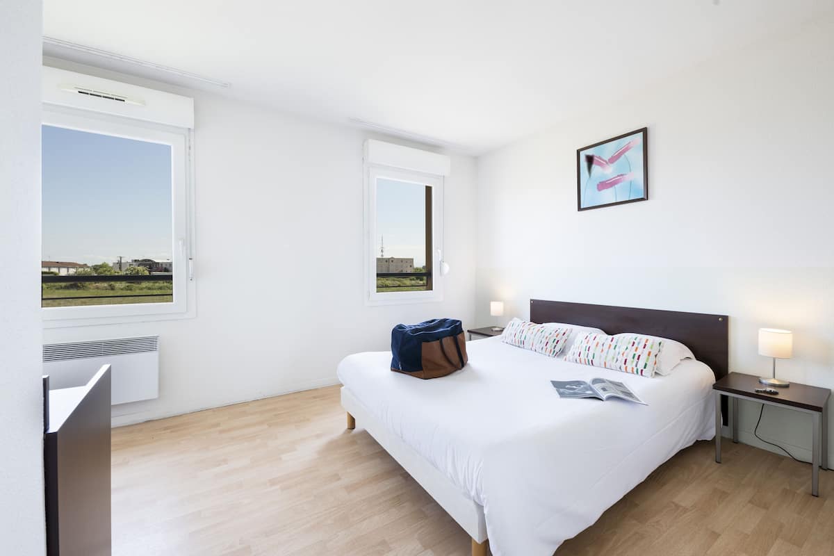 Une chambre d’hôtel moderne avec un grand lit, une table de chevet avec une lampe, une fenêtre donnant sur la ville et un tableau encadré au mur.