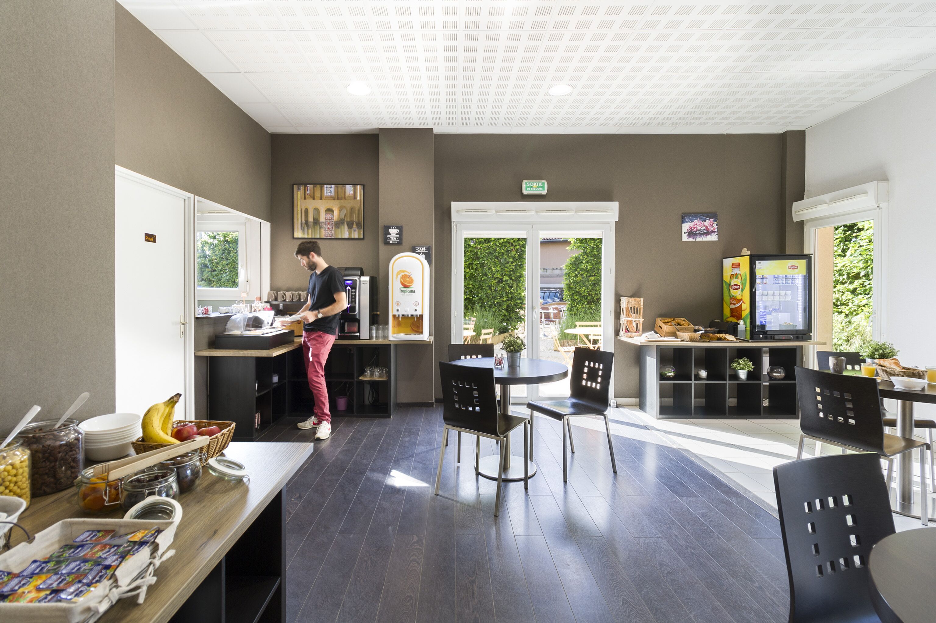 Un espace cuisine moderne où une personne prépare des aliments, un coin repas avec des chaises et des tables, et un réfrigérateur rempli de boissons.