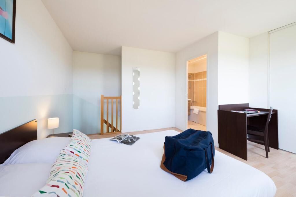 Une chambre d’hôtel avec un lit, un bureau, une chaise et une salle de bain visible à travers une porte ouverte.
