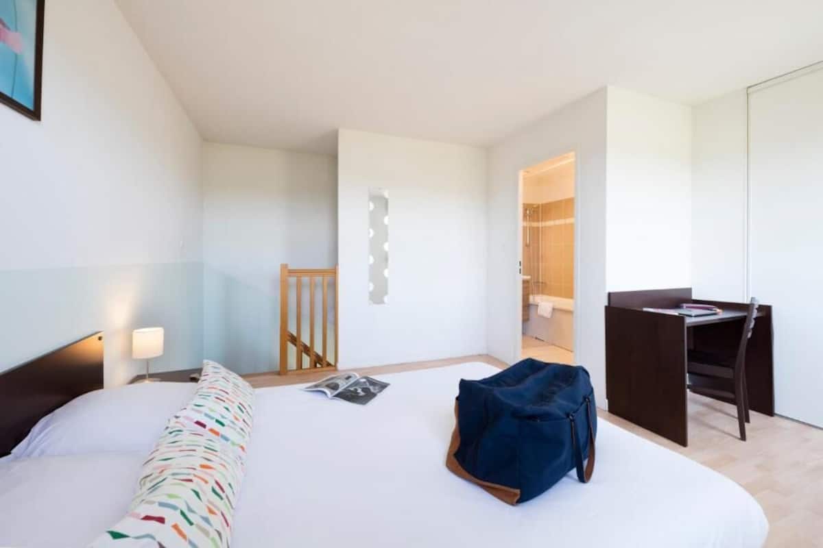 Une chambre d’hôtel avec un lit, un bureau, une chaise et une salle de bain visible à travers une porte ouverte.