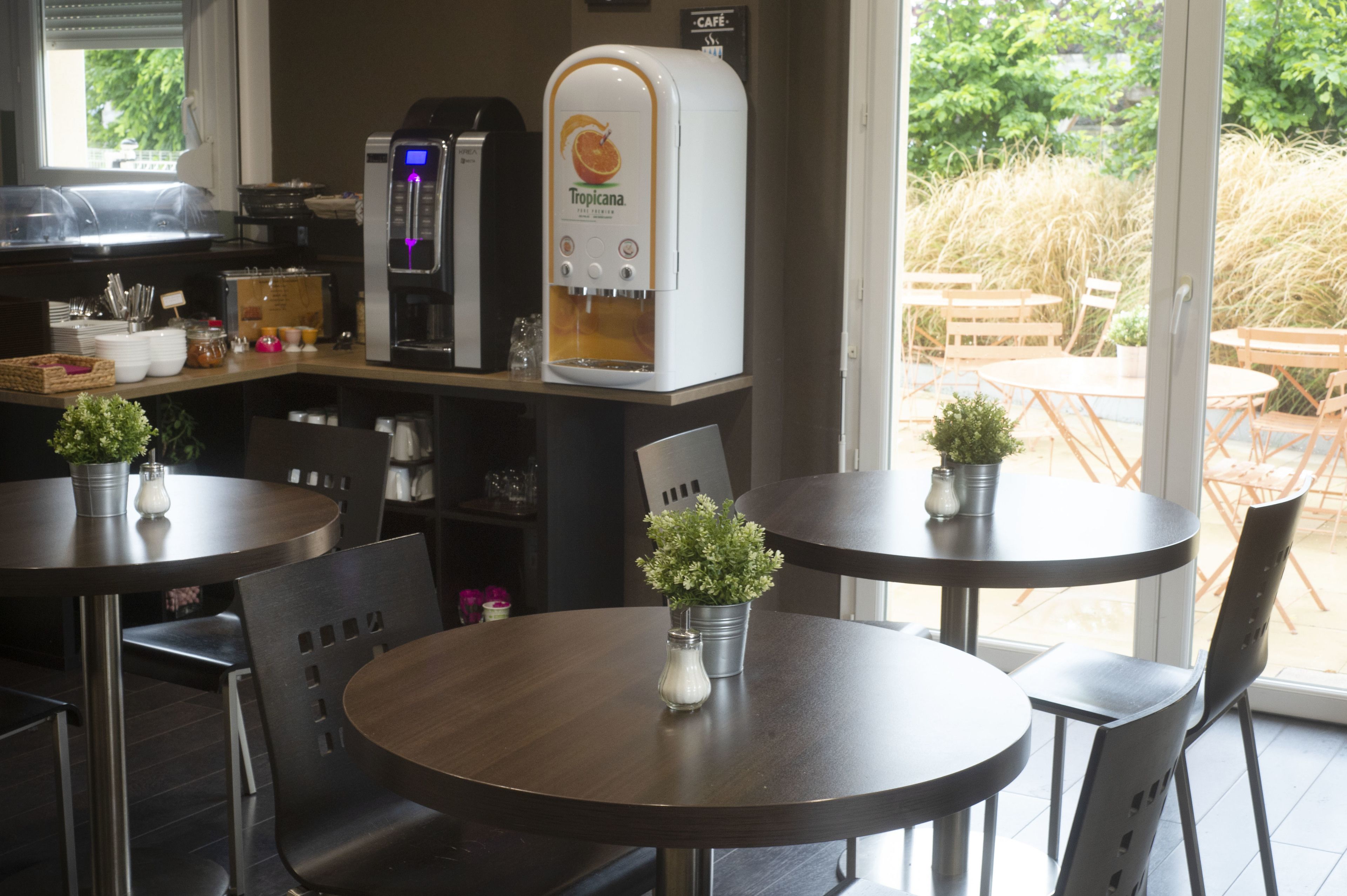 Interior de una cafetería con mesas redondas, sillas, una máquina de café y vistas al área de mesas exteriores.