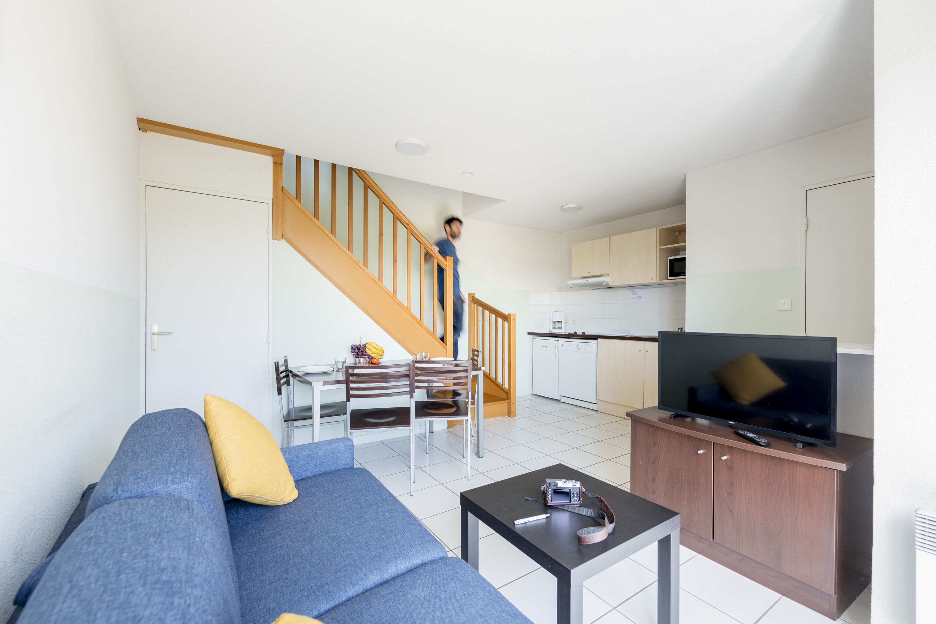 Una sala moderna con sofá, mesa de centro y televisor sobre un mueble de madera. Una escalera conduce a una zona de cocina con armarios y un microondas.