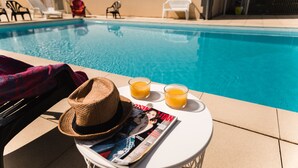 A poolside setting with a magazine, two glasses of orange juice, and a straw hat on a small table.