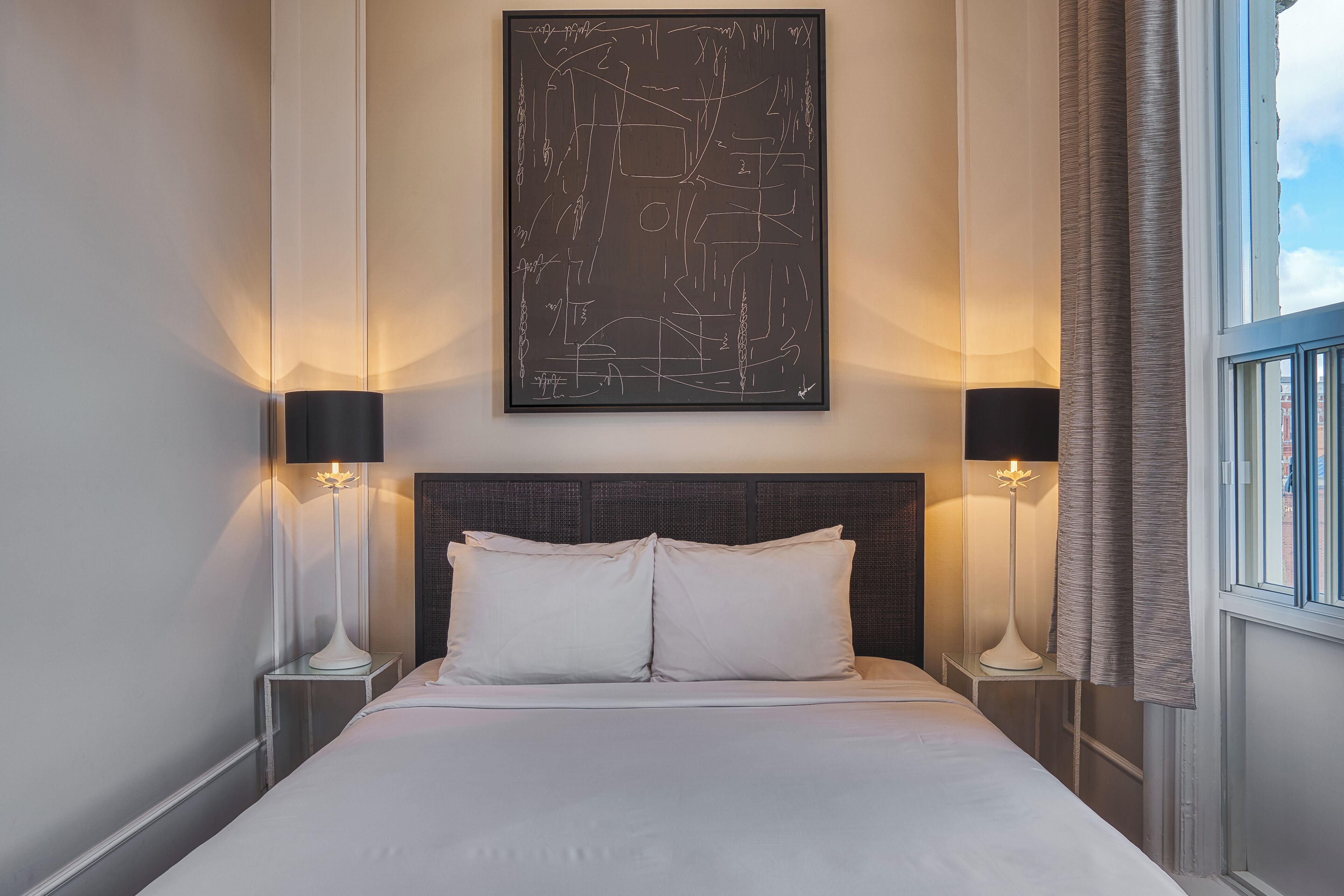 A hotel room with a large bed, two bedside tables, a modern abstract painting, and a window with curtains.