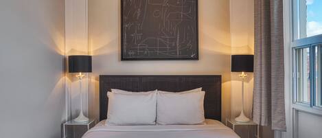A hotel room with a large bed, two bedside tables, a modern abstract painting, and a window with curtains.
