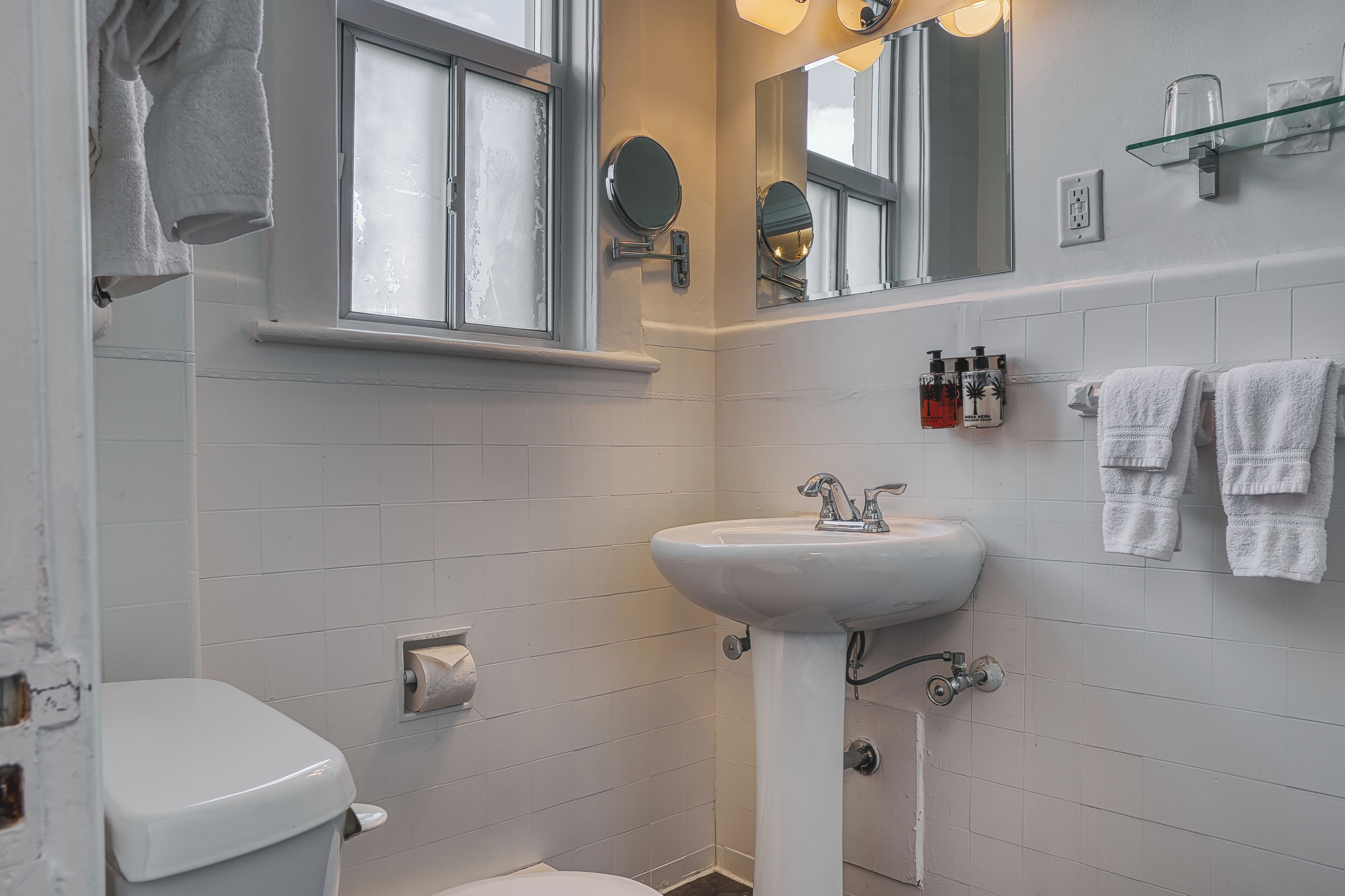 A bathroom with a toilet, sink, and two mirrors. There are towels hanging on the wall and a window with a towel hanging on the outside.