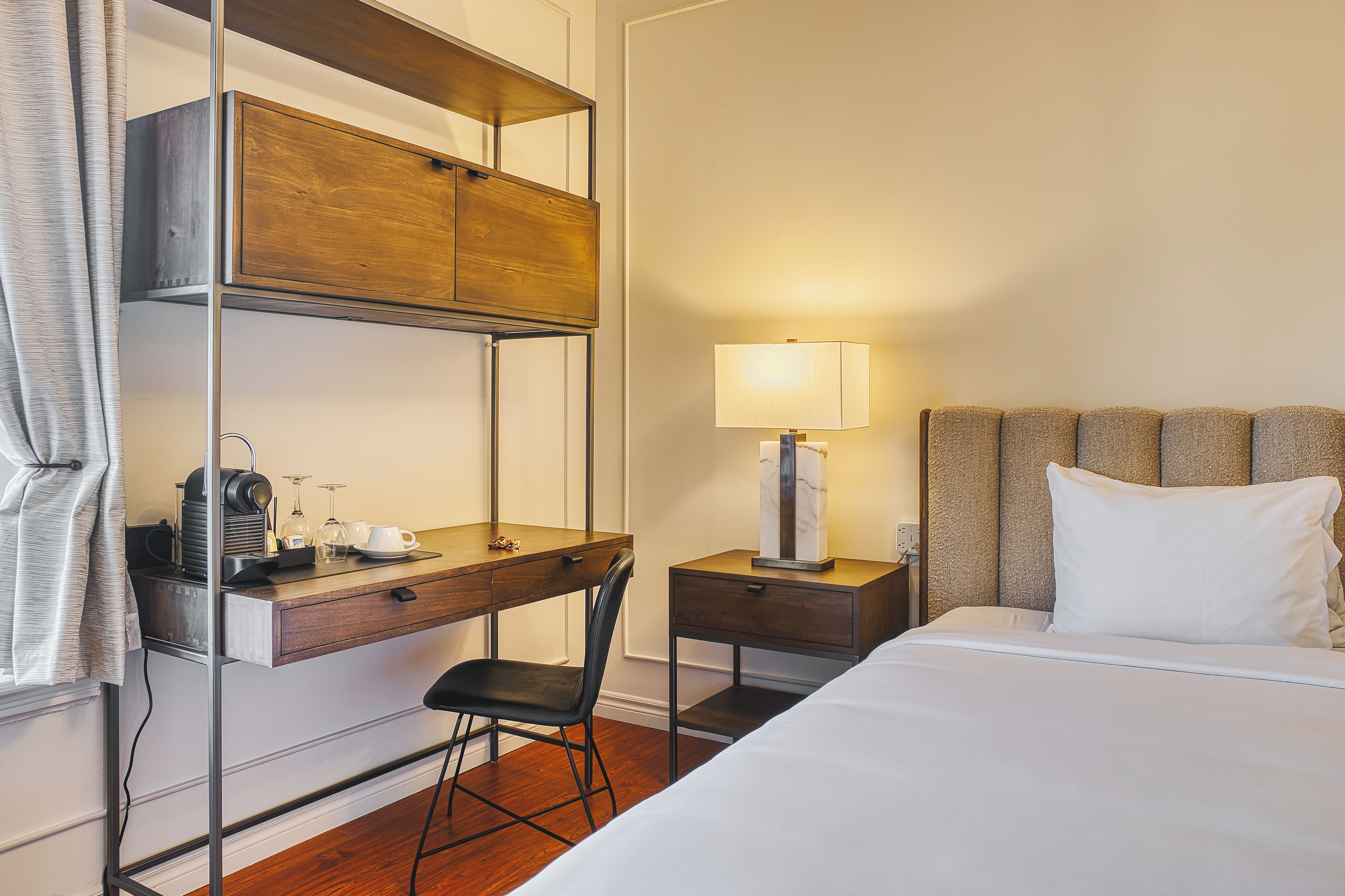A hotel room with a bed, a desk with a chair, a nightstand with a lamp, and a wardrobe.