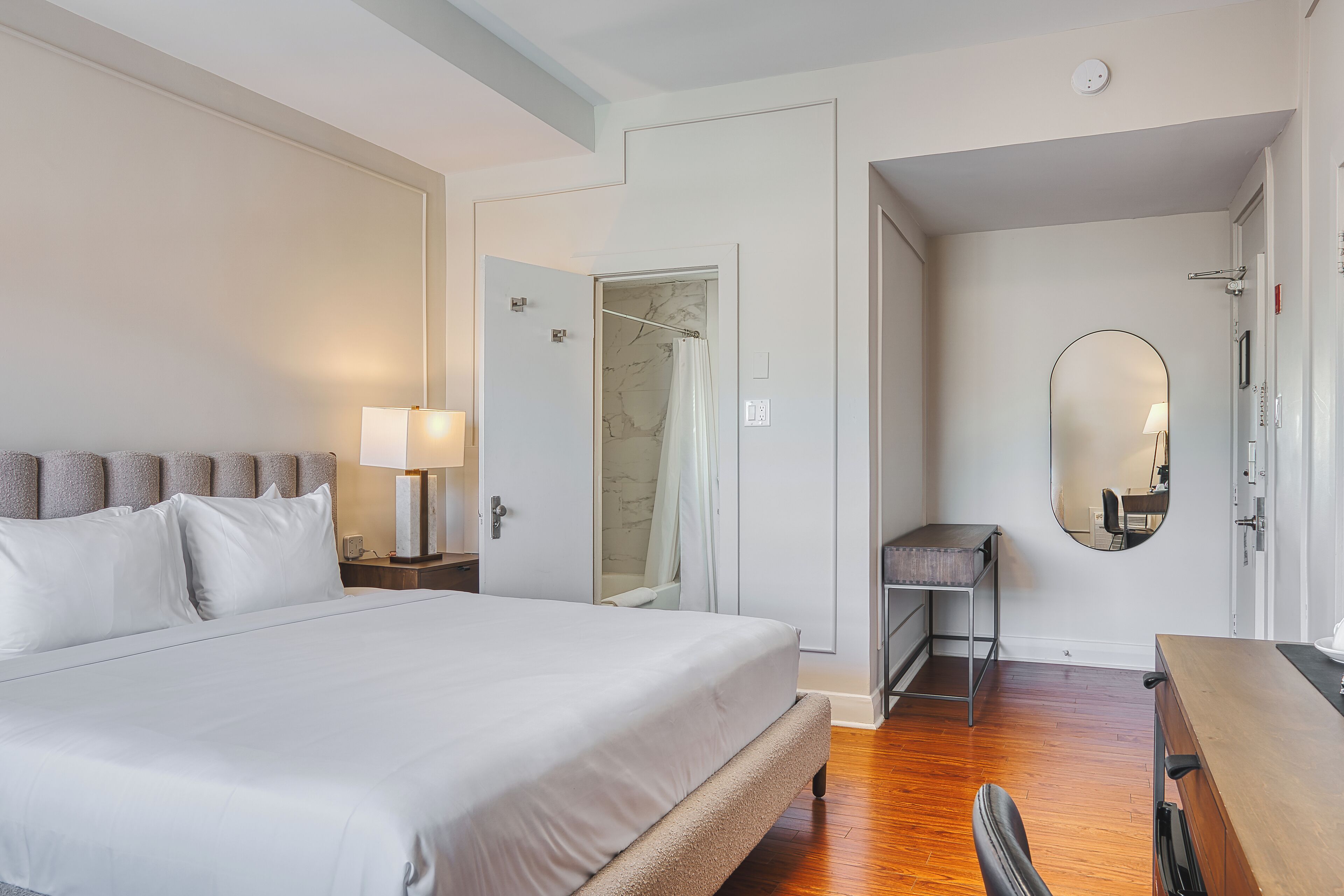 Quarto de hotel com uma cama grande, uma escrivaninha, uma cadeira, um espelho e área de chuveiro.