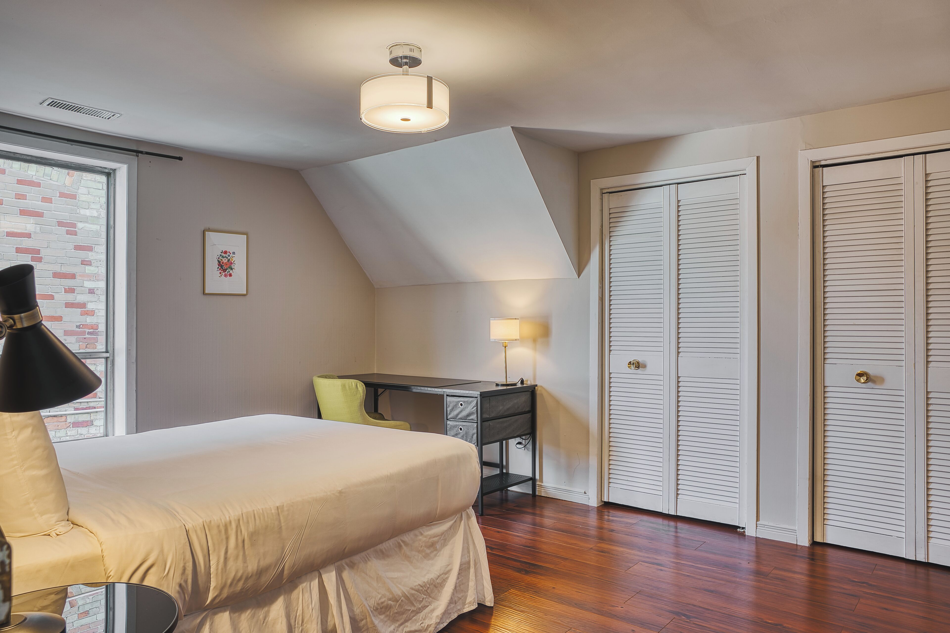 Un dormitorio con cama, escritorio y lámpara. Hay una ventana con vista a un muro de ladrillo y un cuadro en la pared.