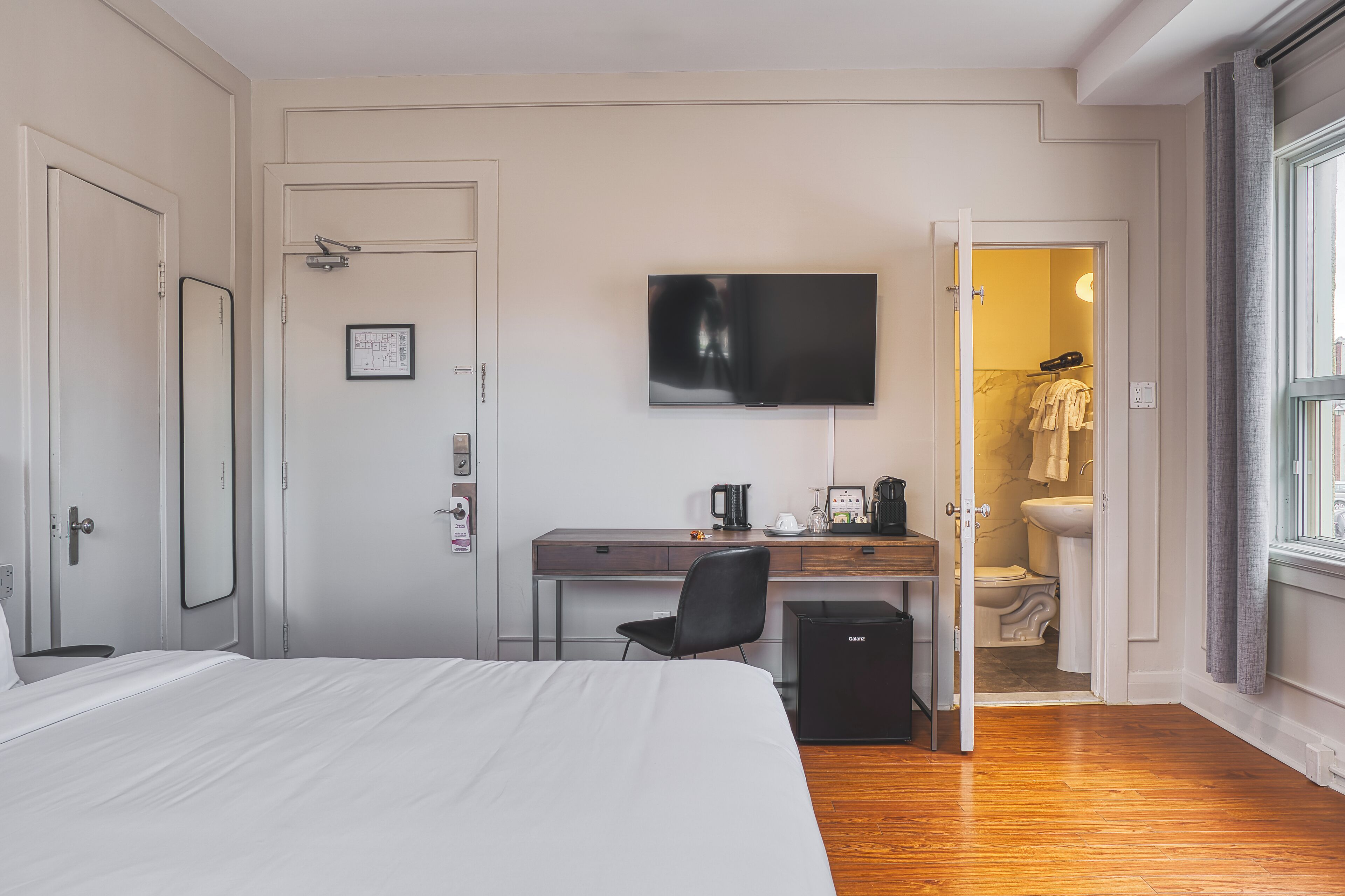 A hotel room with a bed, a desk with a computer, a television, and a bathroom visible through an open door.