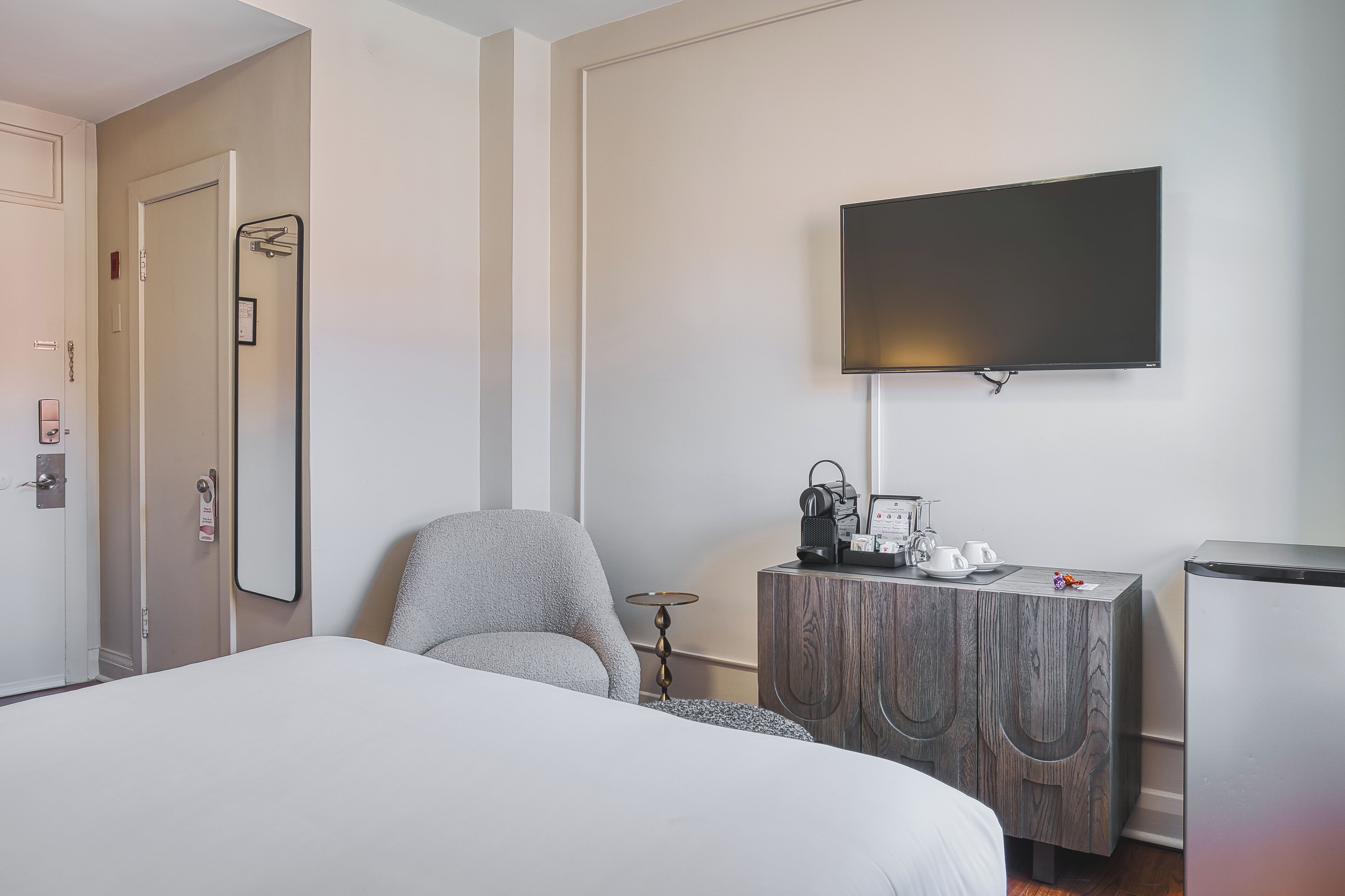 A hotel room with a bed, a grey armchair, a wooden dresser with a flat-screen TV mounted above it, a small table with a lamp, and a minibar.