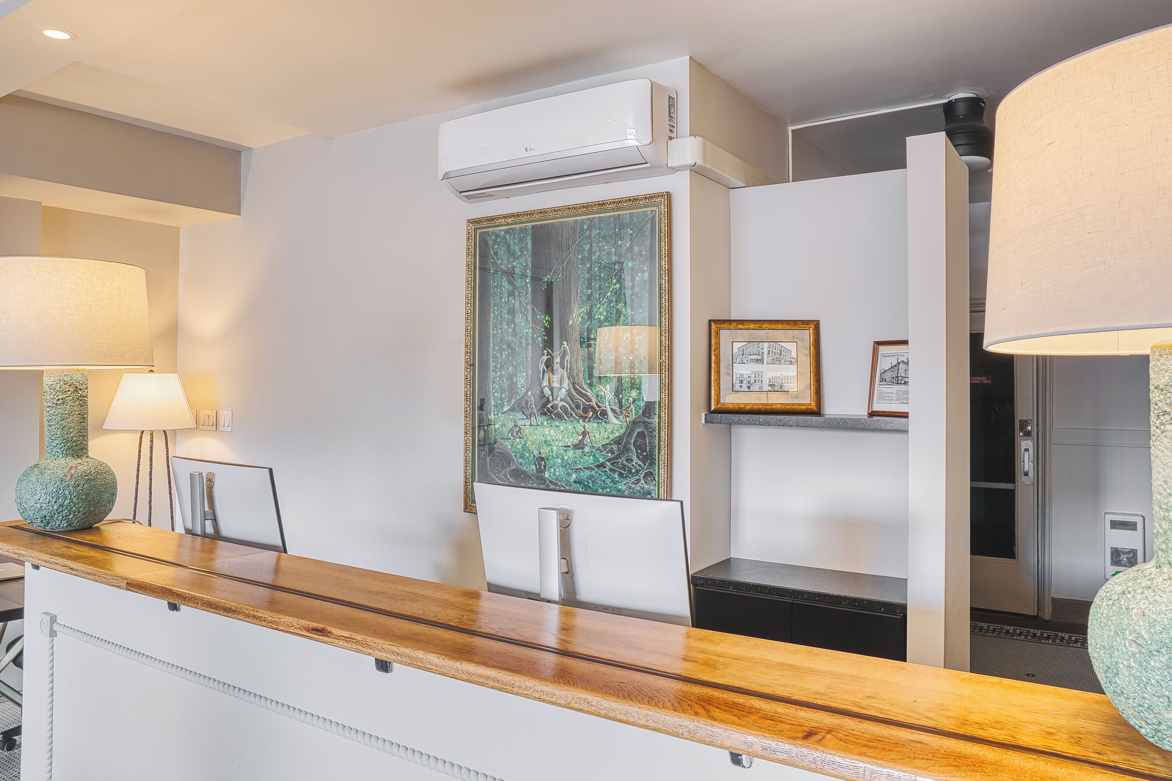 En receptionsdisk med en träbeklädd skiva, en inramad målning som föreställer en skogsmiljö och en vit luftkonditioneringsenhet monterad ovanför.