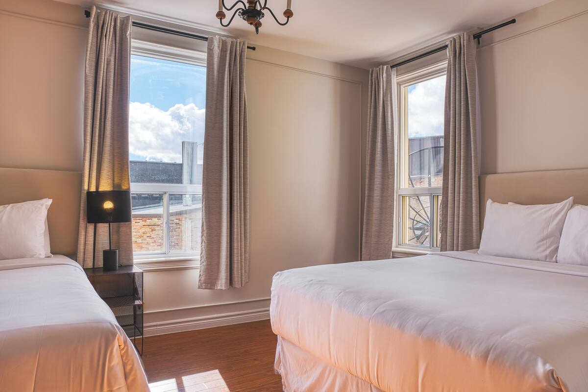 Een hotelkamer met twee bedden, een nachtkastje met een lamp en grote ramen voorzien van gordijnen.