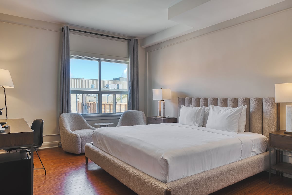 Een hotelkamer met een groot bed, twee stoelen, een bureau en een raam met uitzicht op gebouwen.