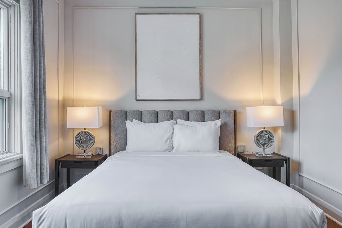 Een net opgemaakt bed met witte lakens, twee nachtkastjes met lampen, een groot lijstwerk boven het bed en een raam met gordijnen aan de linkerkant.