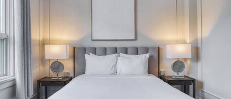 A neatly made bed with white linens, two bedside tables with lamps, a large framed artwork above the bed, and a window with curtains on the left.