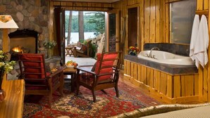 Ein Holzhütten-Interieur mit Kamin aus Stein, einer Holzwanne und Blick auf einen See.
