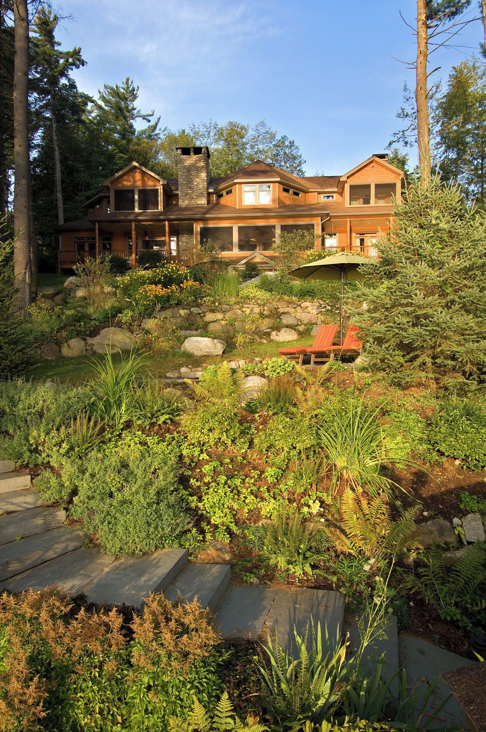A large, multi-story house with a stone chimney, surrounded by a lush garden with various plants and a stone pathway leading to a red lounge chair.