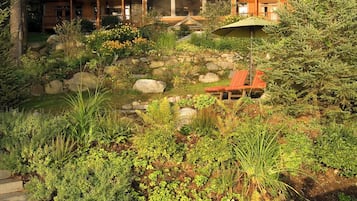 A large, multi-story house with a stone chimney, surrounded by a lush garden with various plants and a stone pathway leading to a red lounge chair.